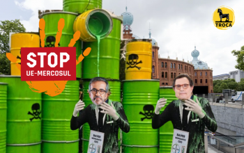 Imagem de teatro: vários barris a verterem líquido verde atrás de actores com máscara dos ministros.